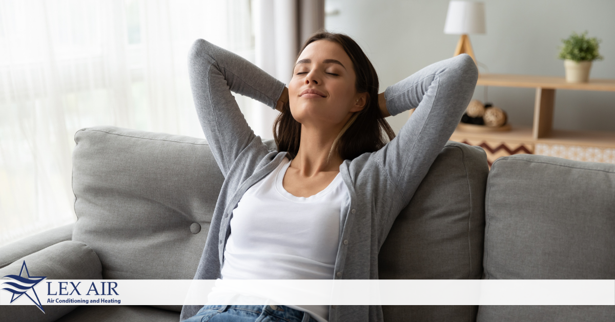 A woman with her eyes closed, smiling, with her hands behind her head sitting on a gray couch