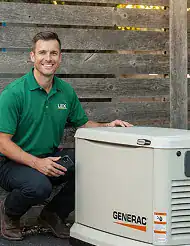 A man in a green polo shirt kneels next to a Generac generator with a wooden fence in the background.