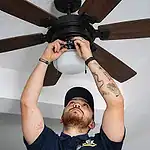 %Imagetitle% | Lex Cooling, Heating Plumbing &Amp; Electrical A person wearing a blue shirt and cap stands on the floor and fixes the wiring of a ceiling fan with a light fixture.