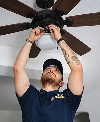 A person wearing a blue shirt and cap stands on the floor and fixes the wiring of a ceiling fan with a light fixture.