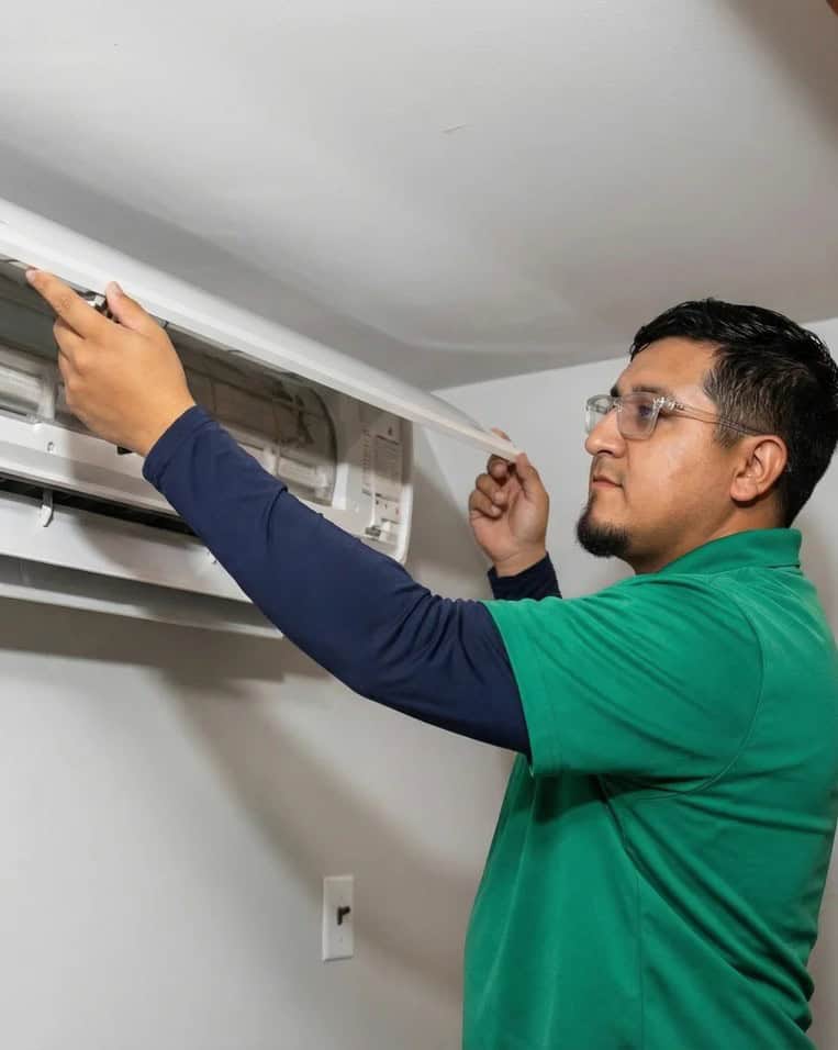 A man in a green shirt and glasses is inspecting or repairing a wall-mounted air conditioning unit indoors.