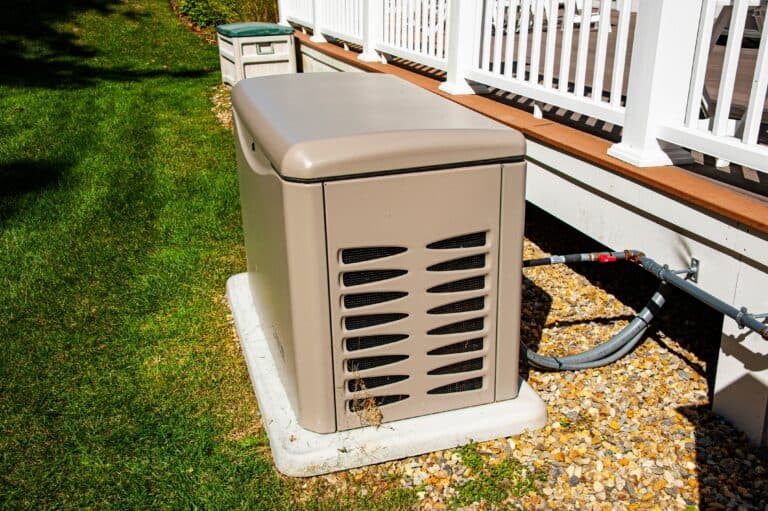 A beige backup generator sits on a concrete pad next to a house, connected by cables and surrounded by grass and gravel.
