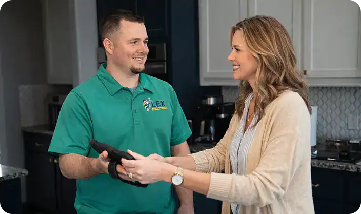 A man in a green polo shirt hands a black device to a smiling woman in a kitchen.