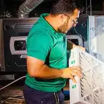 %Imagetitle% | Lex Cooling, Heating Plumbing &Amp; Electrical A man wearing glasses and a green shirt replaces an air filter in an HVAC unit inside a mechanical room.