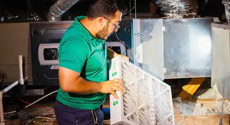 A man wearing glasses and a green shirt replaces an air filter in an HVAC unit inside a mechanical room.