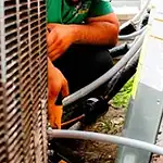 %Imagetitle% | Lex Cooling, Heating Plumbing &Amp; Electrical A man wearing glasses and a green shirt repairs or inspects an outdoor HVAC unit, using tools near electrical cables.