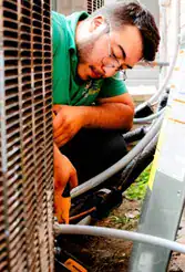 A man wearing glasses and a green shirt repairs or inspects an outdoor HVAC unit, using tools near electrical cables.
