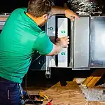 %Imagetitle% | Lex Cooling, Heating Plumbing &Amp; Electrical A person in a green shirt works on an HVAC unit, adjusting a panel with tools and wiring visible nearby.