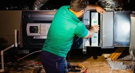 A person in a green shirt works on an HVAC unit, adjusting a panel with tools and wiring visible nearby.