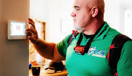 A man in a green work shirt adjusts a wall-mounted digital thermostat set to 70 degrees.